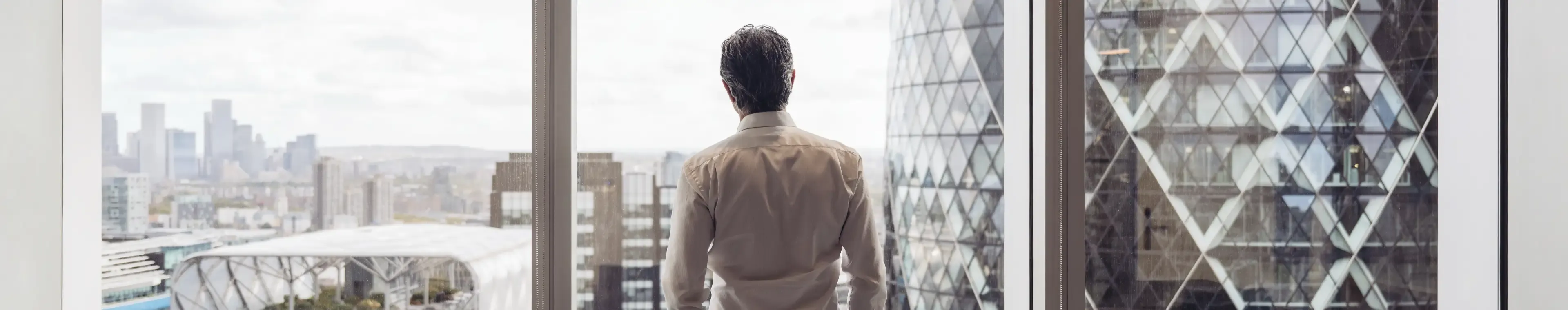 Rear view across conference table in modern board room of executive in early 40s standing with hands in pockets and looking at London’s financial district.