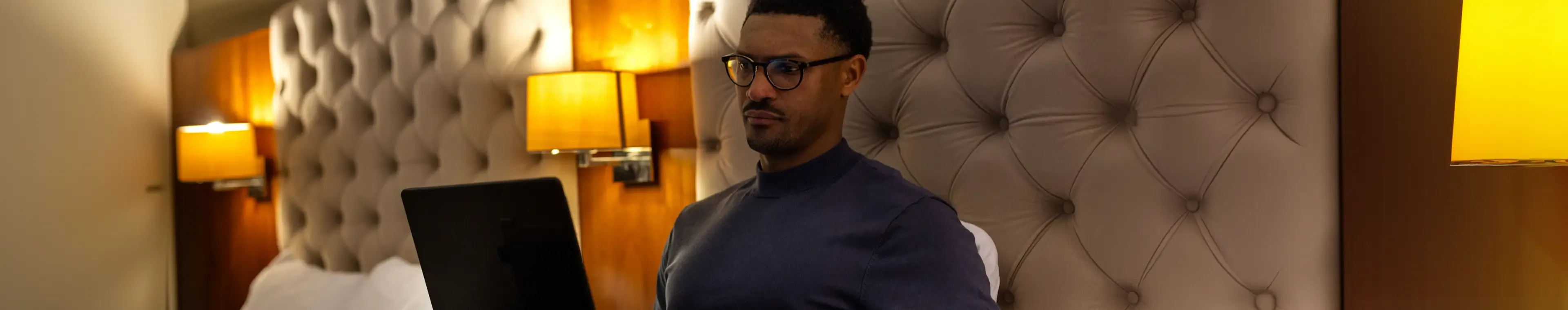 A shot of a mid-adult male sitting on a bed at nighttime in a hotel room in Newcastle Upon Tyne, North East England. He has a laptop in front of him as he works remotely by lamplight whilst away from home. He has a serious expression and is wearing glasses and casual clothing.