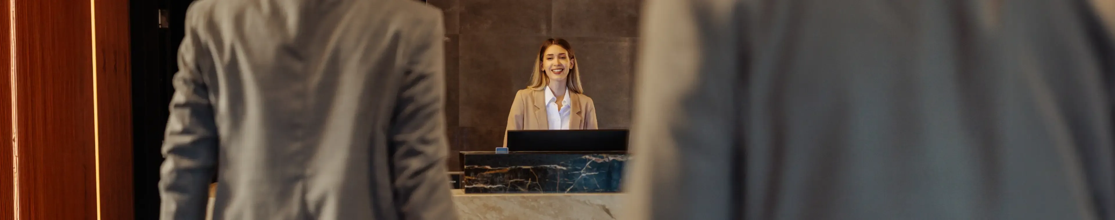 Two suited men walking in the hotel and being welcomed by a young Caucasian receptionist
