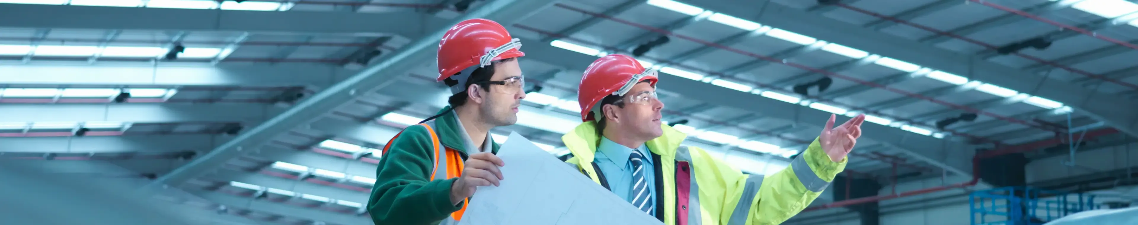 Site foreman advising apprentice with plans in partly completed factory