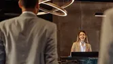 Two suited men walking in the hotel and being welcomed by a young Caucasian receptionist