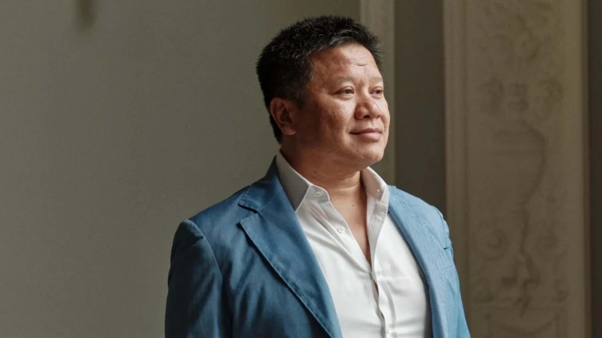 A man in a white shirt and blue blazer with black hair smiling and looking to the side of the camera