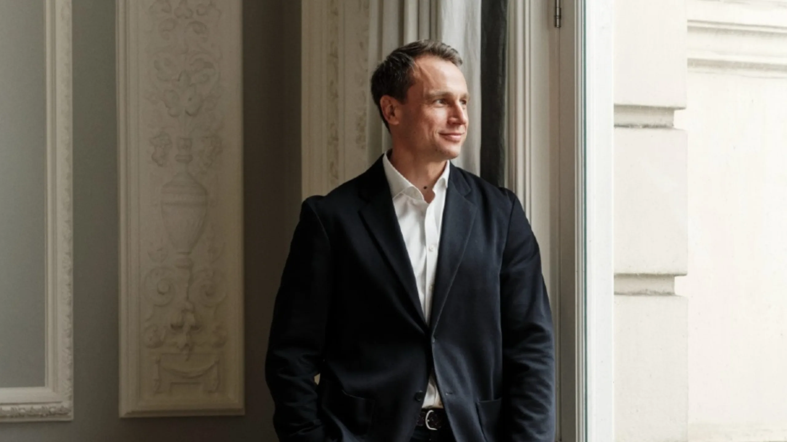 A man stands against a wall looking out the window and smiling dressed in a blue blazer, white shirt and dark blue jeans