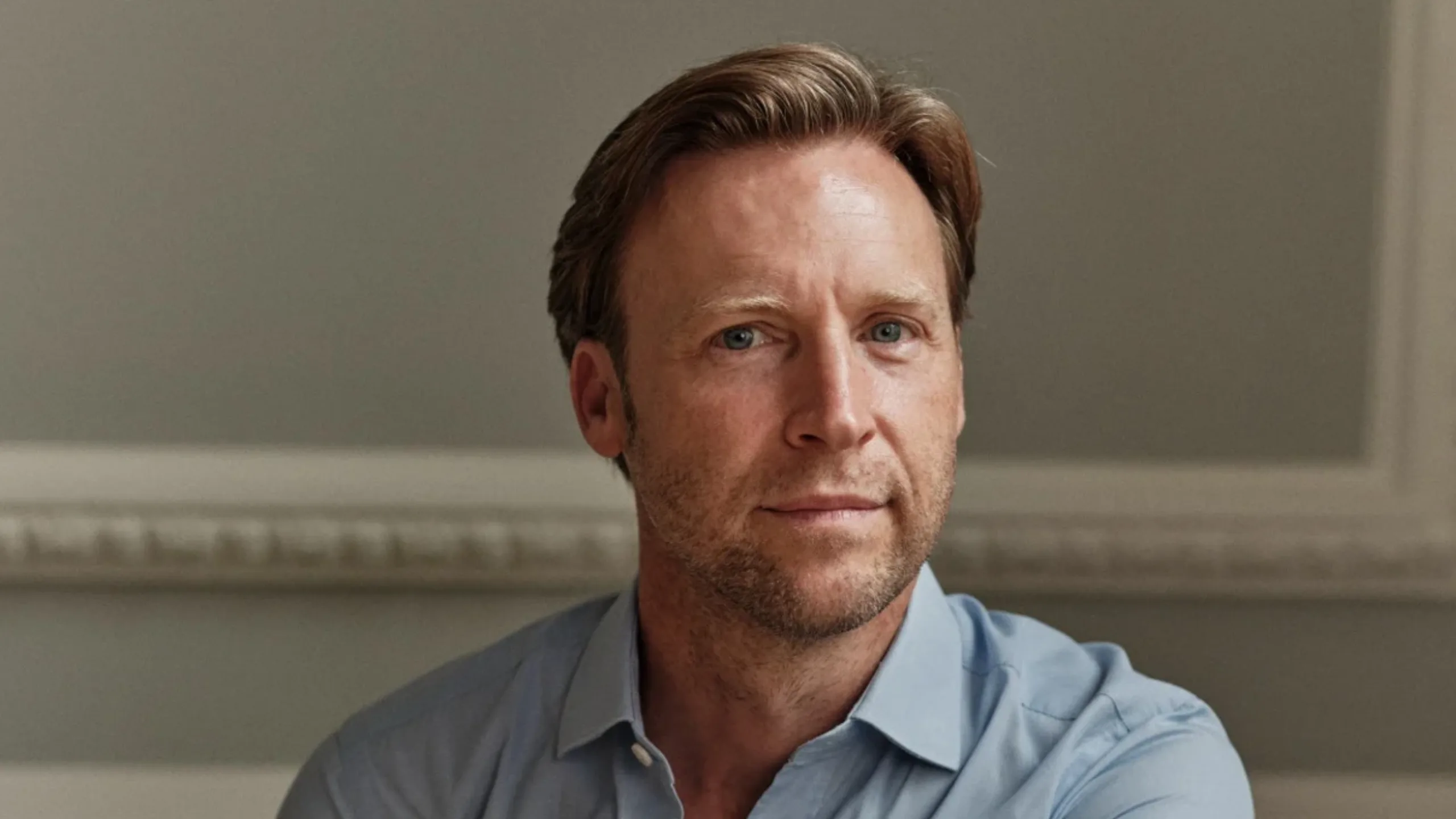 A man in a light blue shirt and blonde hair looking at the camera and smiling slightly